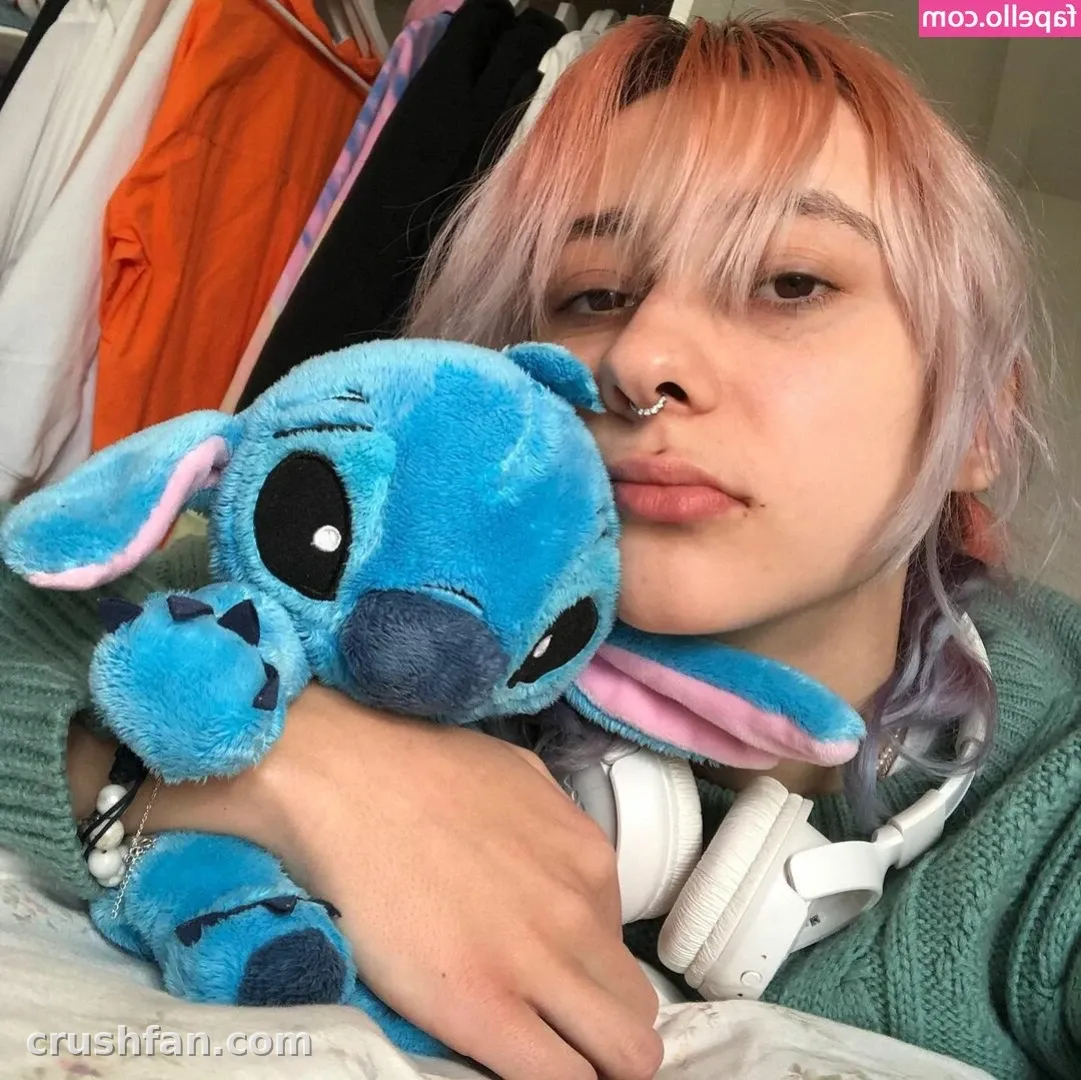 Creator in a green sweater holding a blue plush toy, with white headphones around the neck.