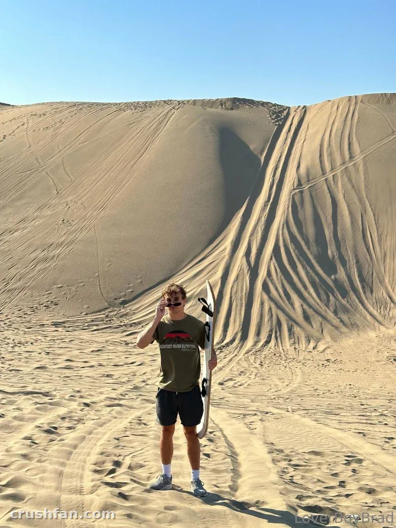 Leo winkt und hält ein Board vor großen Sanddünen unter einem klaren Himmel