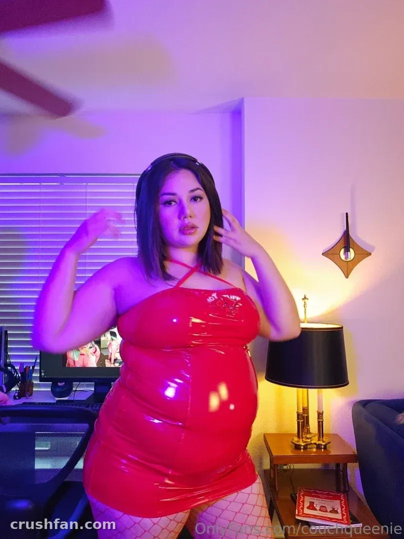 CouchQueen in a shiny red dress posing indoors under purple lighting.