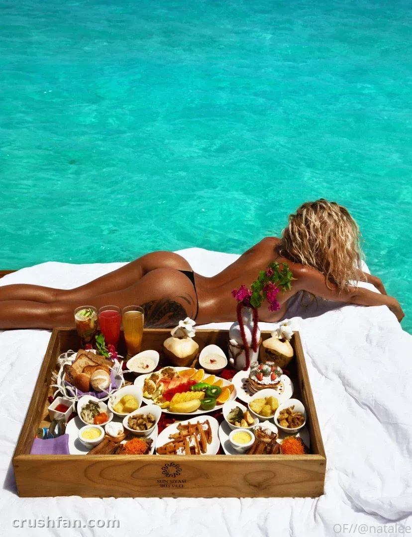 Nata Lee allongée près de l'eau turquoise à côté d'un plateau de petit-déjeuner en bois avec des fruits et du jus.
