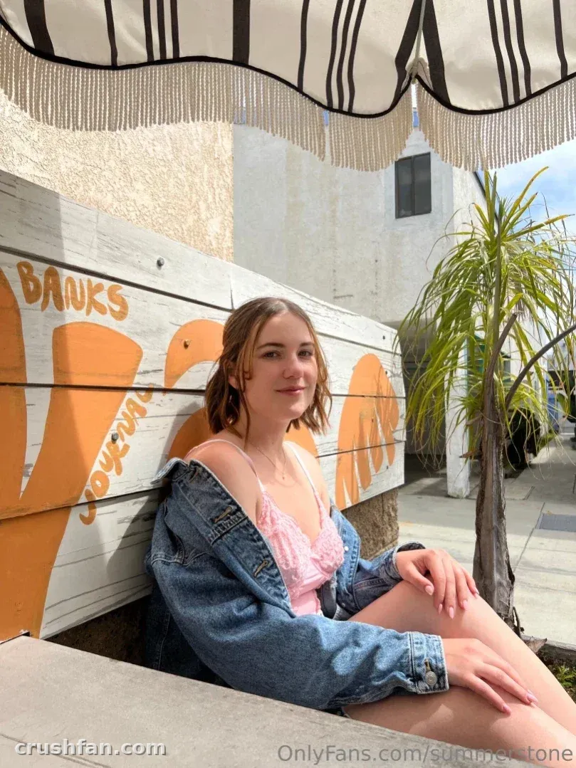 Summer sits outdoors in a denim jacket and pink top beside a painted wooden wall.