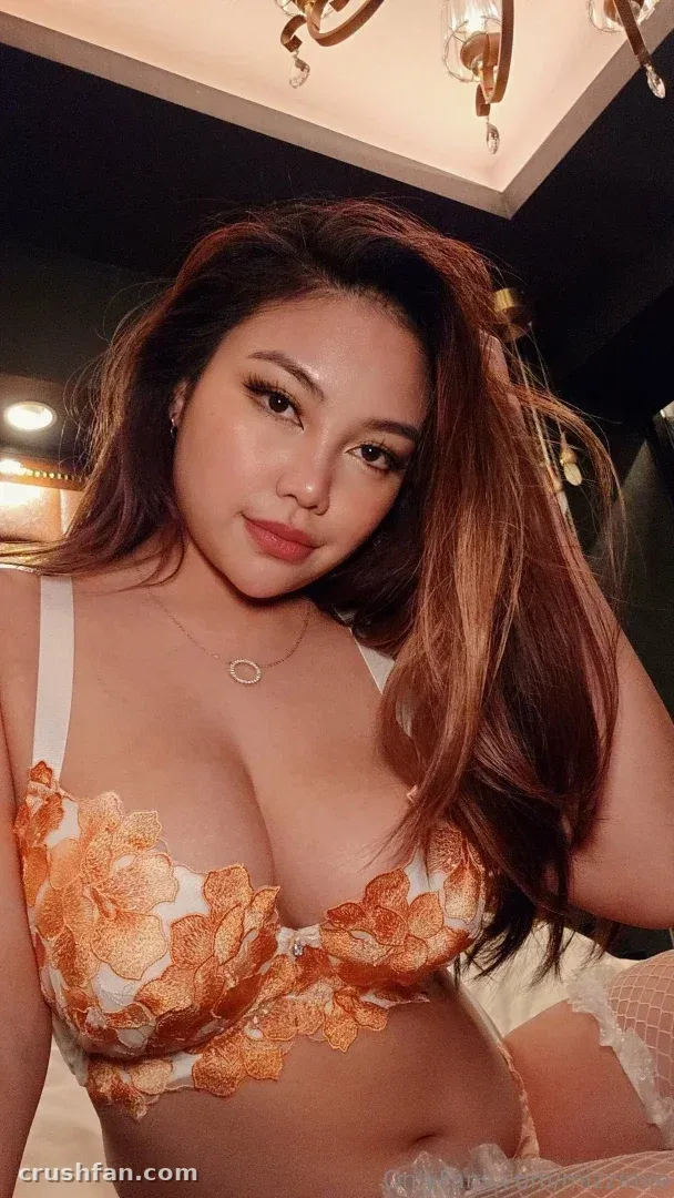 Mizzi Mie in an orange and white floral lace bra, photographed indoors under warm light