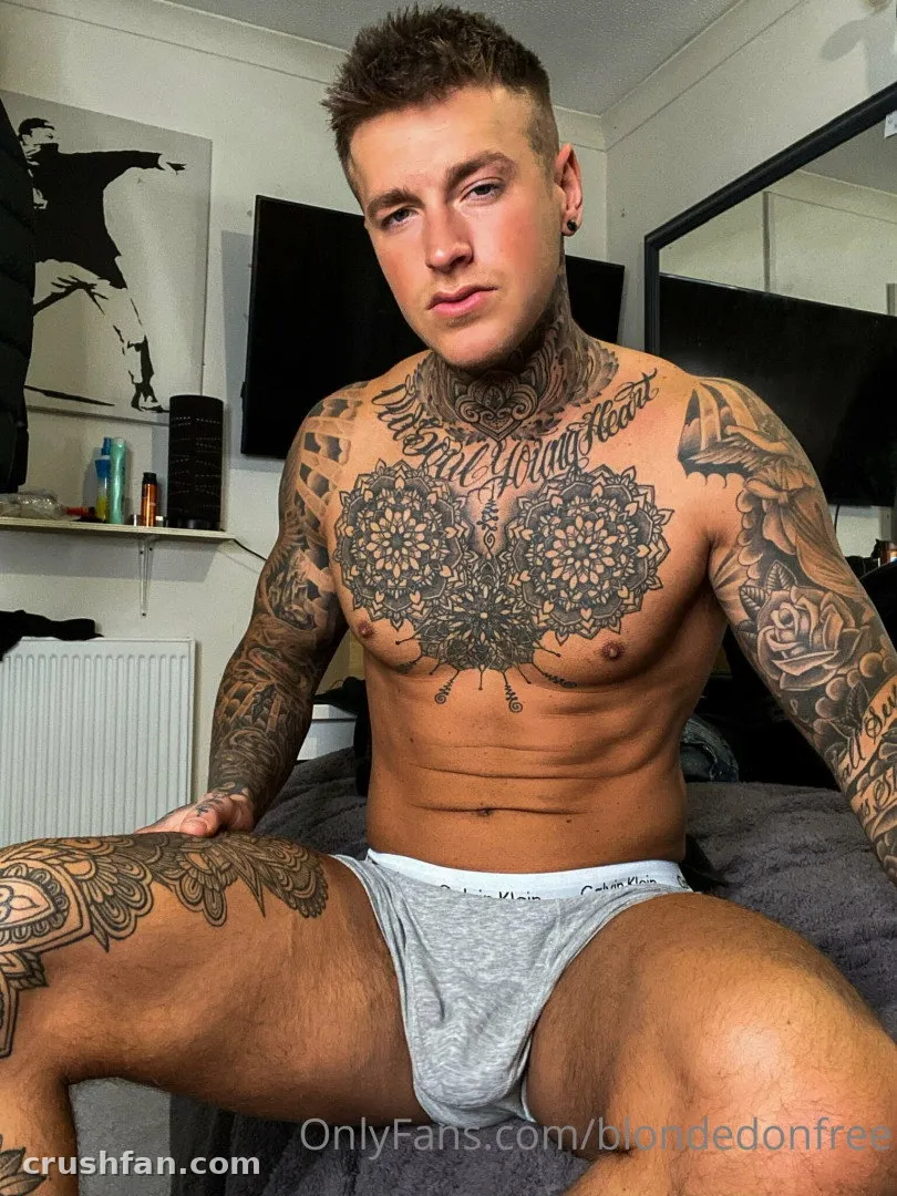 Tätowierter Mann in grauer Unterhose sitzt auf einem Bett in einem sanft beleuchteten Raum
