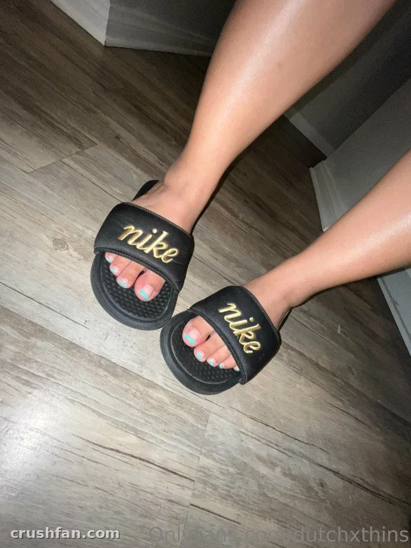 Vue rapprochée de pieds en sandales claquettes noires avec des ongles vert pâle sur un sol en bois gris.
