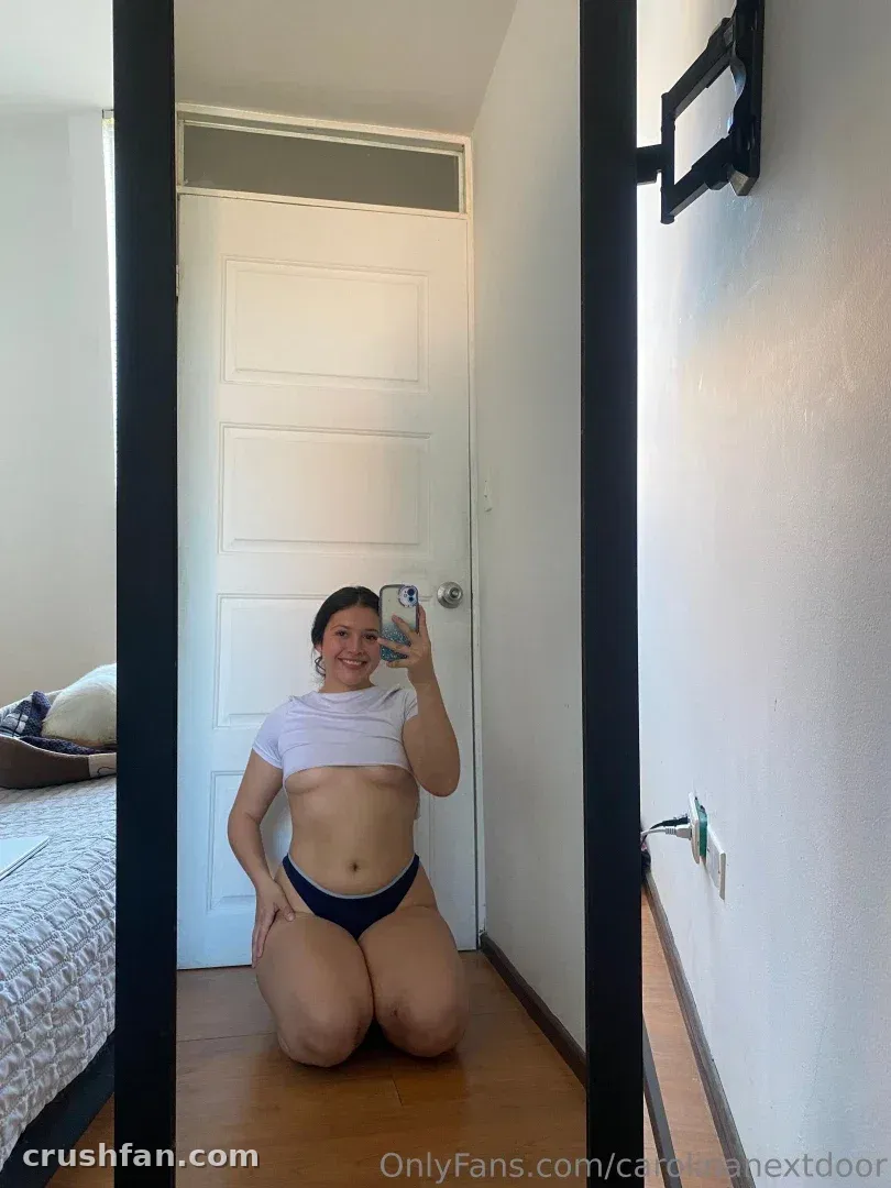 Carolina ajoelhada em um piso de madeira tirando uma selfie no espelho com um top branco e calcinha escura.
