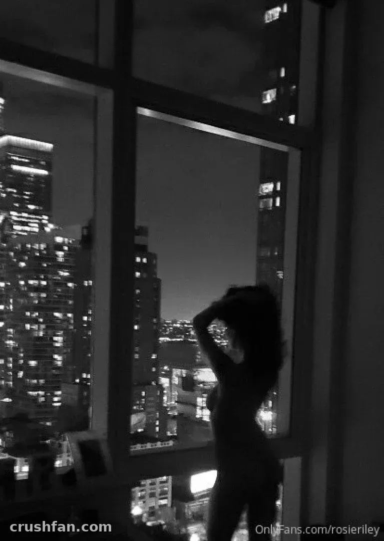 Photo en noir et blanc d'une femme en silhouette à une fenêtre donnant sur une ligne d'horizon urbaine éclairée la nuit