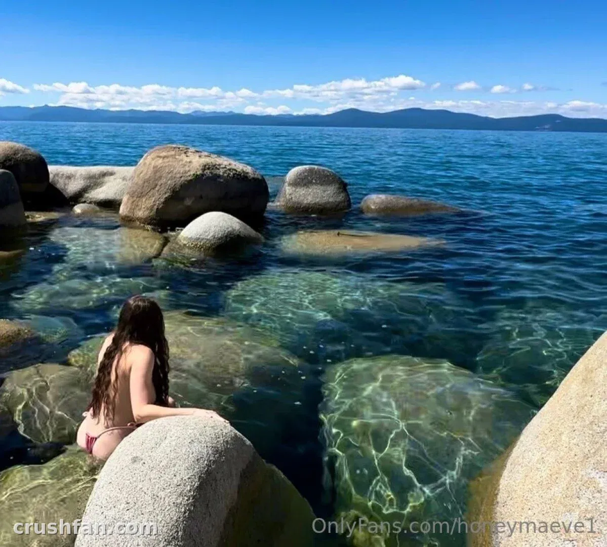 Maeve in a bikini sitting on a rock beside clear blue lake water
