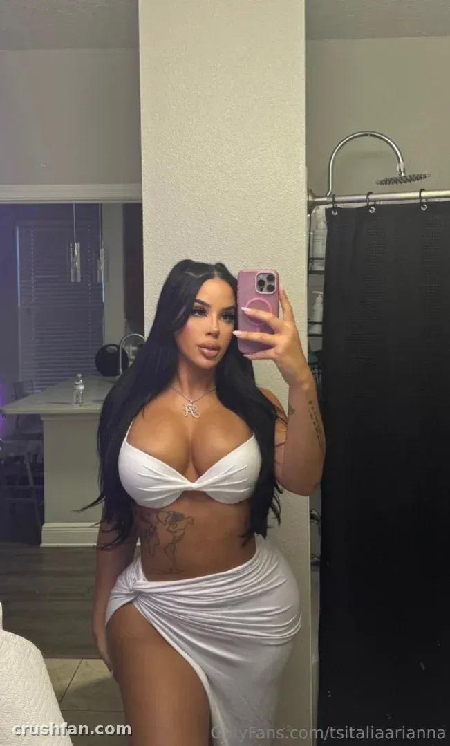 Italia Arianna in a white top and skirt taking a mirror selfie indoors.