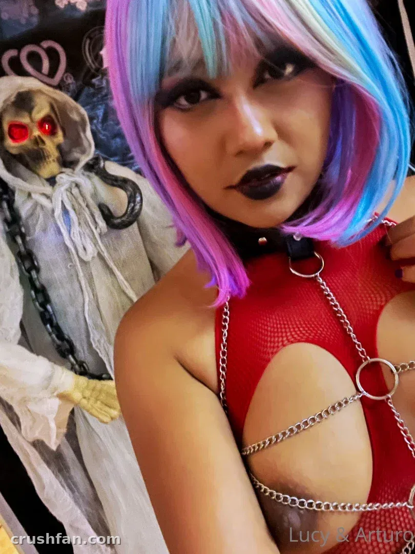 Lucy in a blue and pink wig and red chain-trimmed top posing in front of a reaper decoration