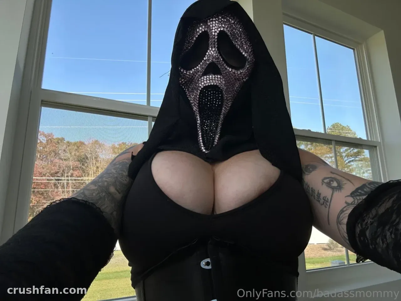 Sara com uma blusa preta com capuz e uma máscara de ghostface com strass posando perto de uma janela