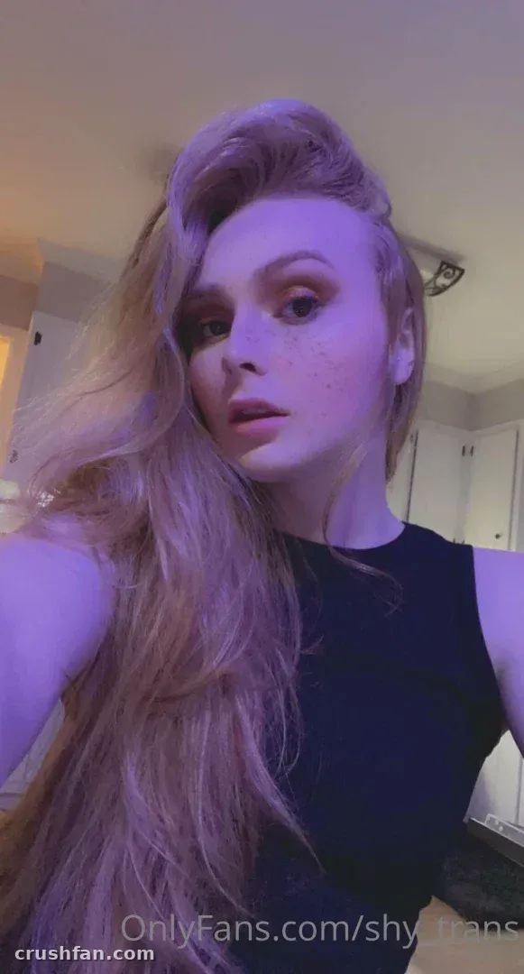 Sarah avec de longs cheveux ondulés et un haut noir, éclairée par une lumière violette en intérieur.