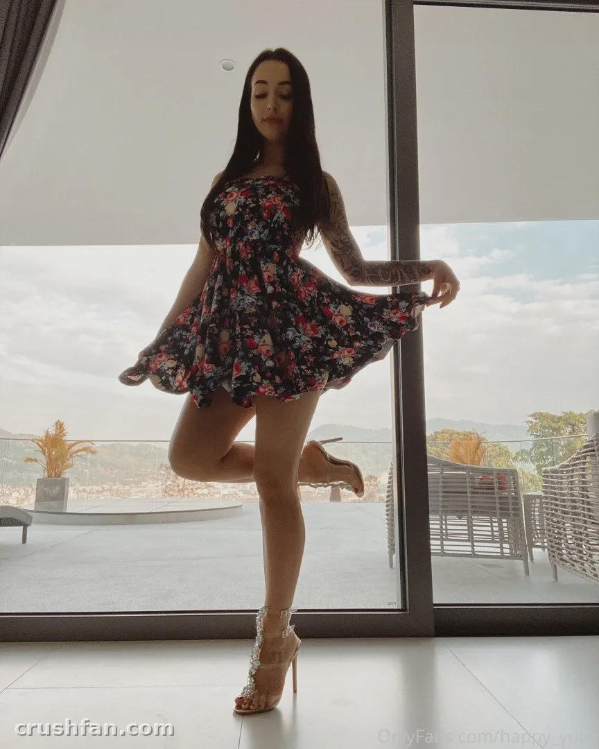 Mujer con un vestido floral oscuro y tacones posando junto a una gran ventana con vistas a una terraza