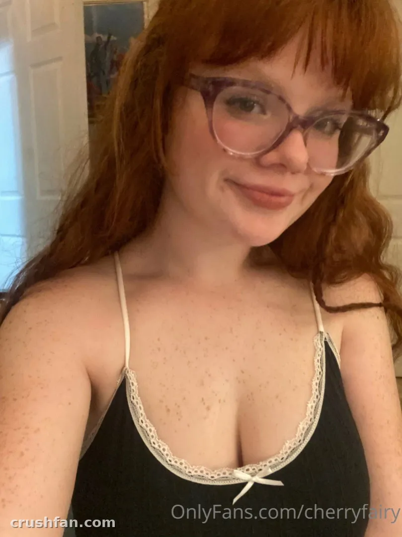 Cherry com uma camisola preta com acabamento de renda e óculos transparentes sorrindo em uma selfie em ambiente interno