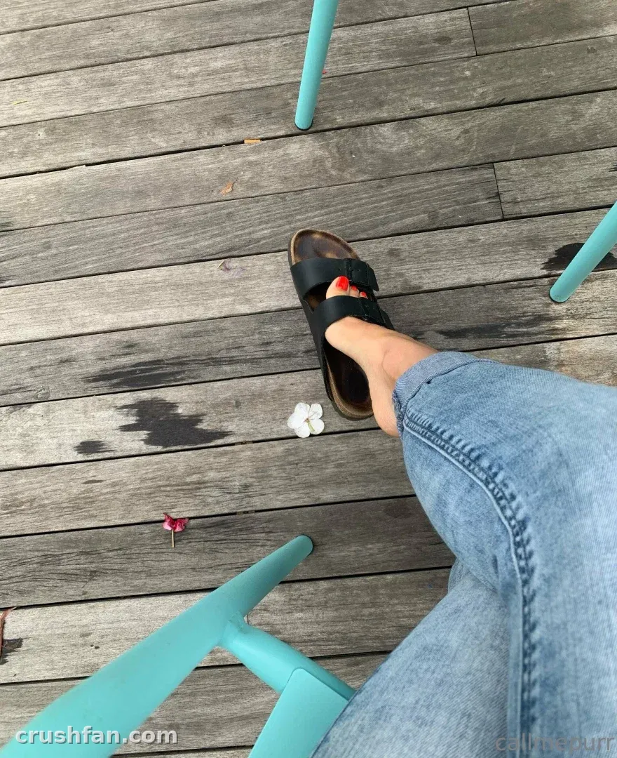Draufsicht auf einen Fuß in einer schwarzen Slide-Sandale und blauer Jeans auf einer Holzterrasse.