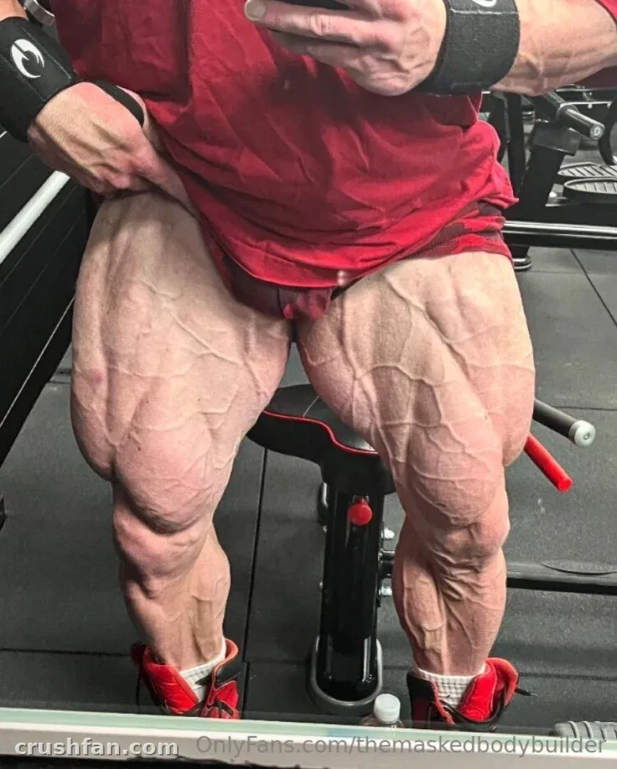 Muscular legs of a bodybuilder in a red shirt seated on a gym bench taking a mirror selfie