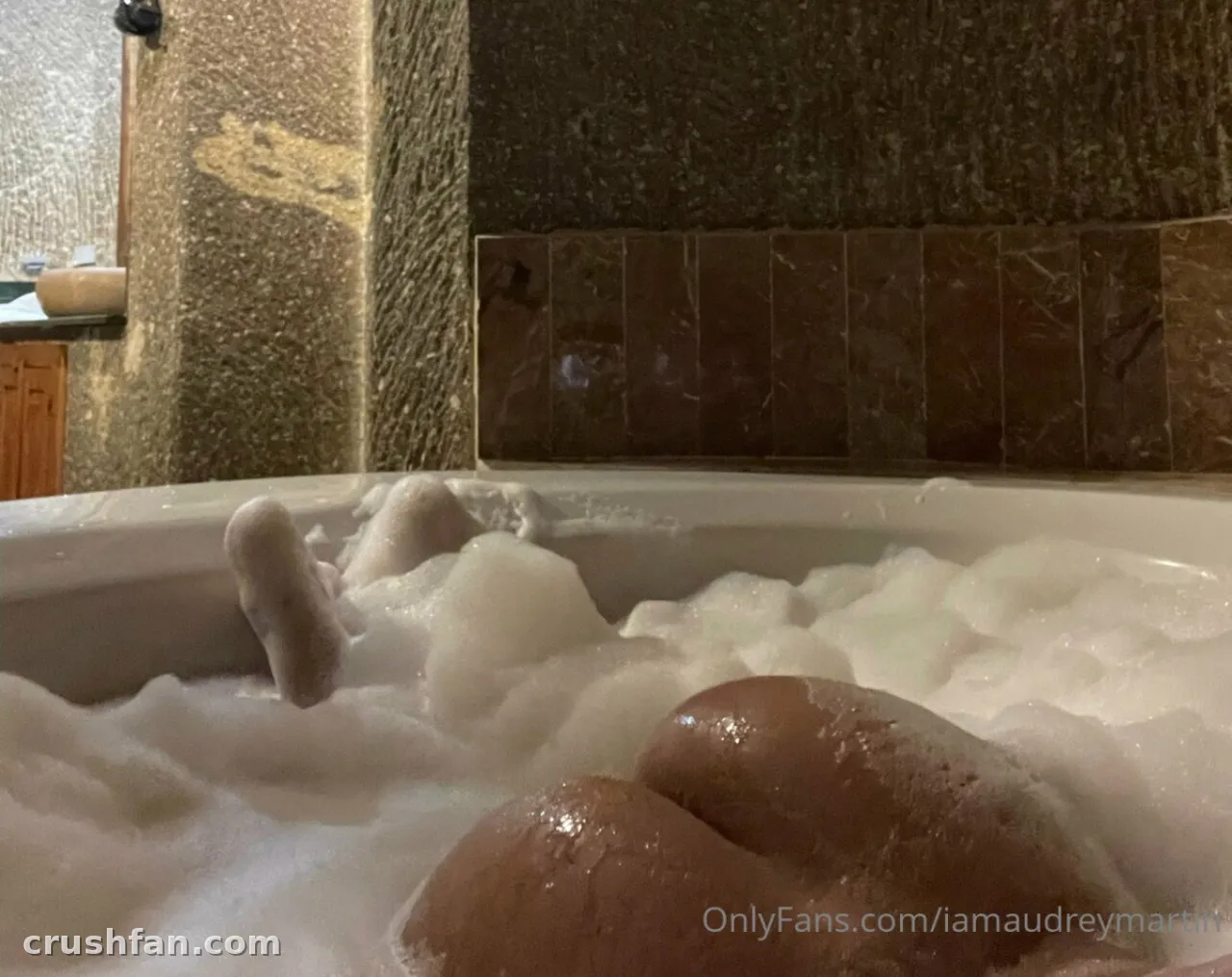 Audrey Martin relaxing in a bubble-filled bathtub with her legs raised, in a warm bathroom setting.