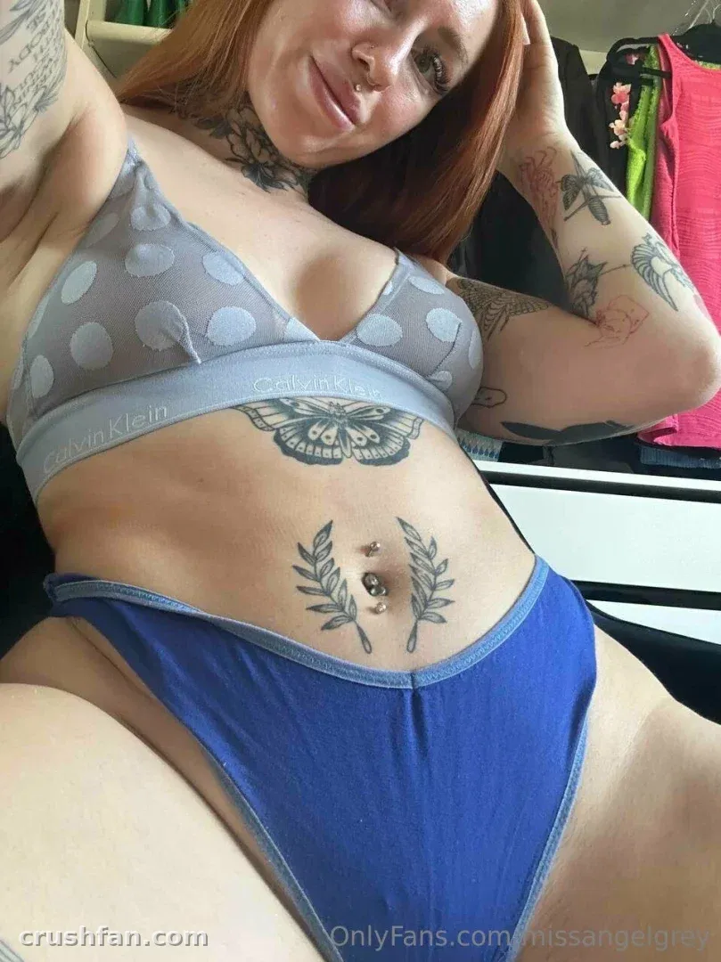 Missangelgrey con sujetador gris de lunares y bragas azules, recostada en interiores con tatuajes visibles.
