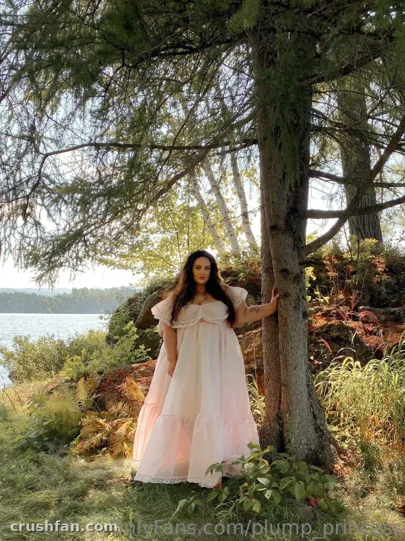 Creator dans une robe rose pâle debout près d'un arbre au bord d'un lac