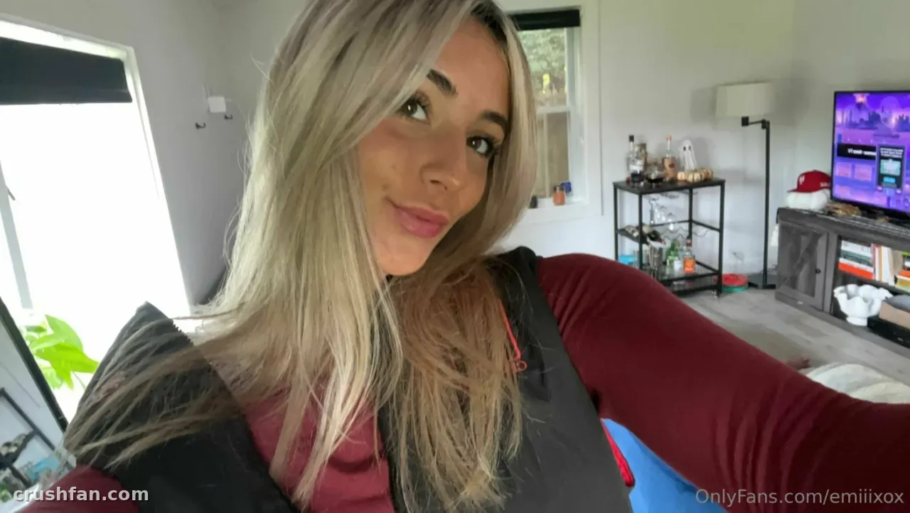 Em smiling in a close-up selfie with long blonde hair, wearing a dark jacket over a red top indoors.