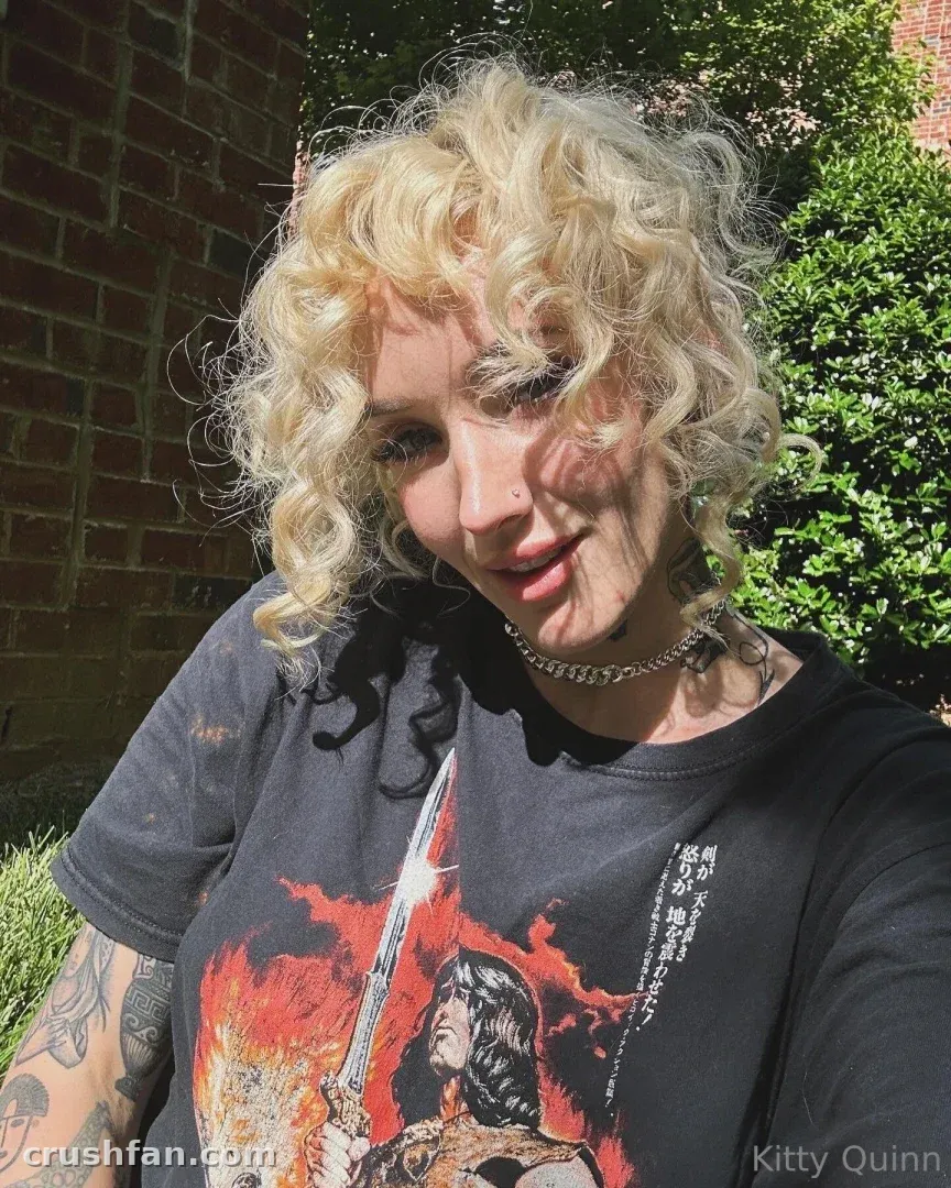Too Twisted Taboo VIP tomándose una selfie al sol al aire libre con una camiseta negra gráfica y cabello rubio rizado.