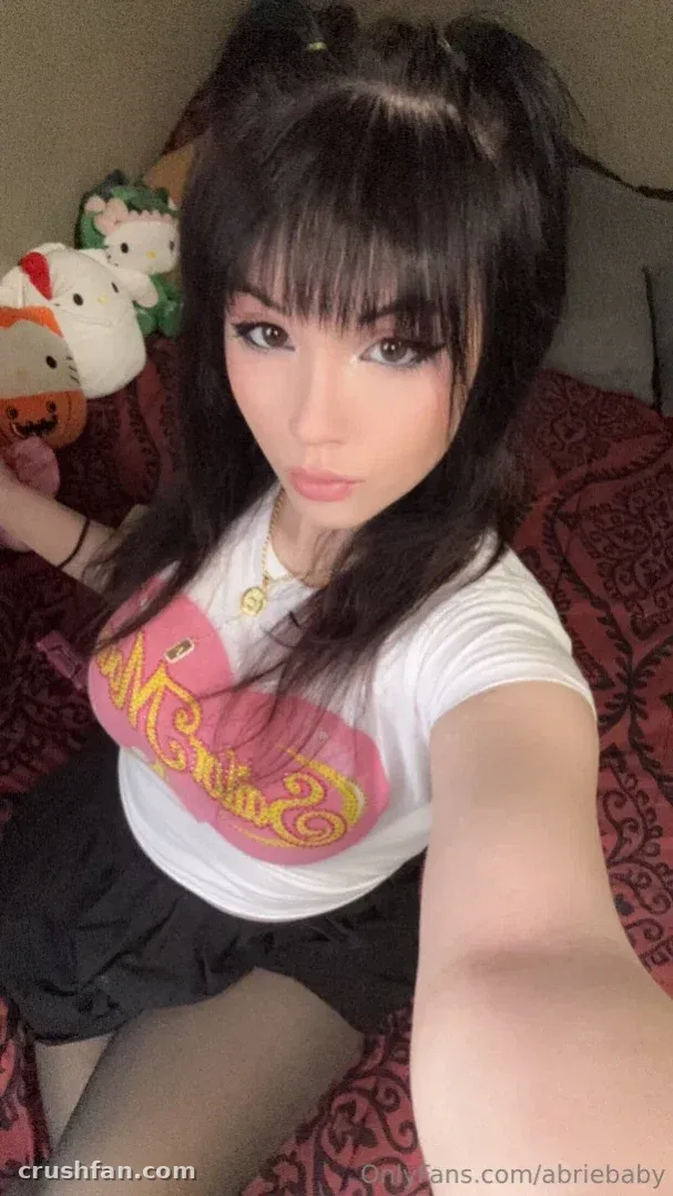 Abriebaby con una camiseta gráfica blanca y una falda oscura tomándose una selfie en un dormitorio.