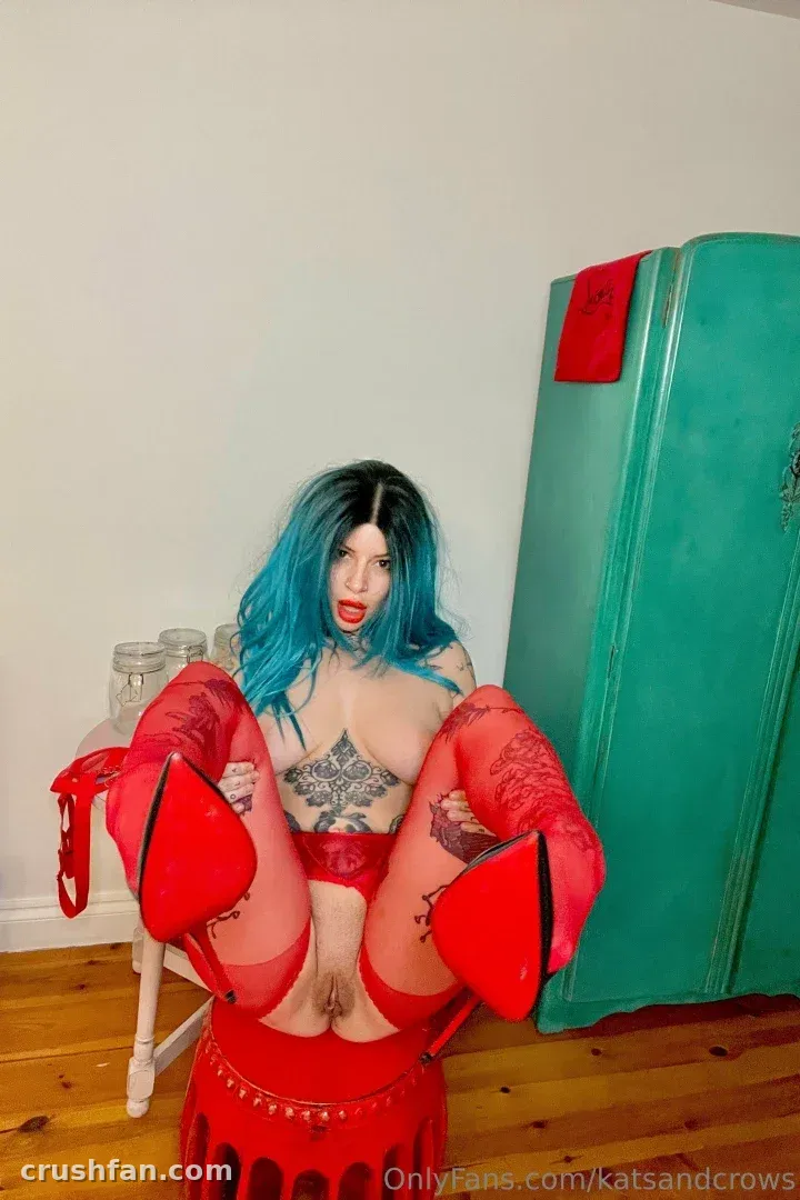 Woman with blue and black hair in red lingerie and red heels posing indoors beside a teal cabinet