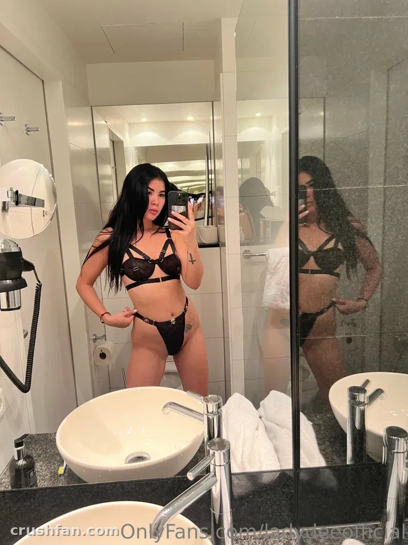Lady Dee de lingerie preta tirando uma selfie no espelho em um banheiro claro