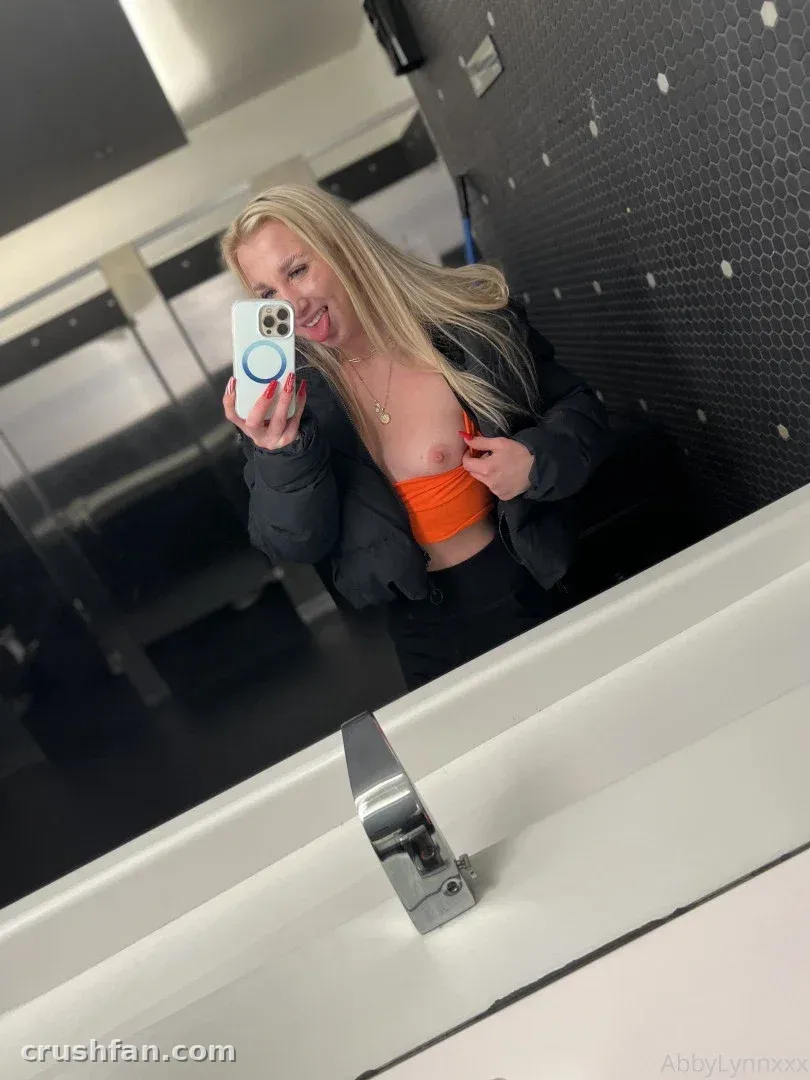 Abby Lynn Fit tirando uma selfie de espelho em um banheiro, usando um top laranja e uma jaqueta escura.