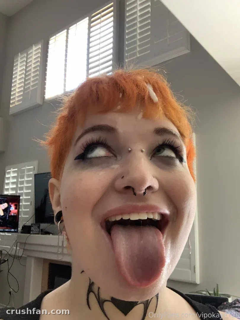 Kaye with short orange hair and a neck tattoo making a playful face indoors.