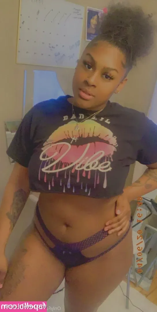 Sweettoothh in einem dunklen grafischen Crop-Top und lila Dessous, drinnen posierend.