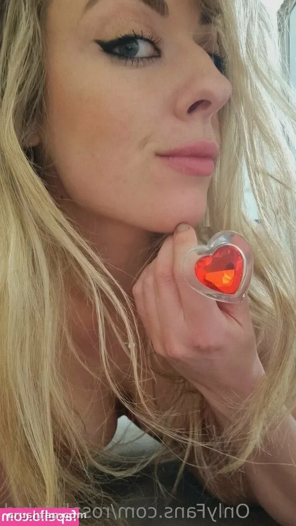 Retrato en primer plano de Rosa Brighid con cabello rubio y un anillo de corazón rojo luminoso cerca de su barbilla