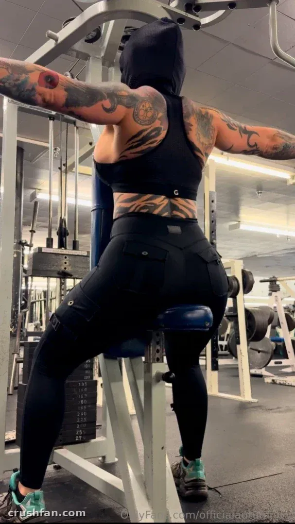 Autumn Ivy in black gym clothing using a weight machine, tattoos visible on her arms