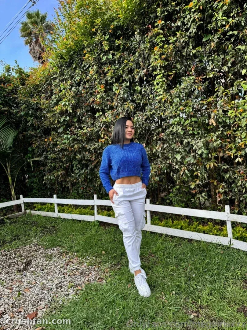 Megan Iris in einem blauen Strickoberteil und grauer Hose, die an einem weißen Zaun in einem grünen Garten steht.