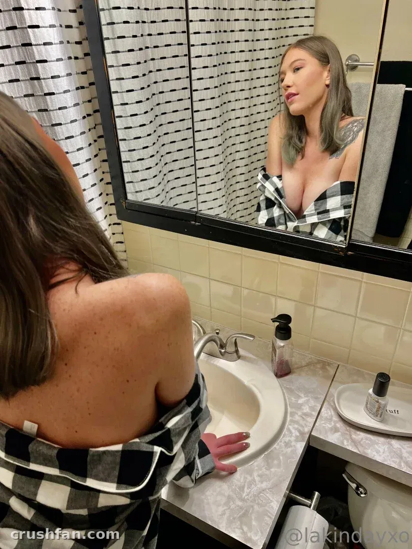 Créatrice en chemise à carreaux penchée au-dessus d'un lavabo de salle de bain, son reflet visible dans le miroir