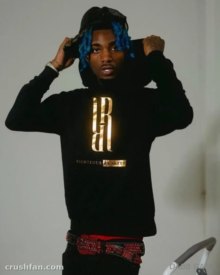 DABB LAND in a black hoodie with a gold emblem, holding the hood with both hands indoors.