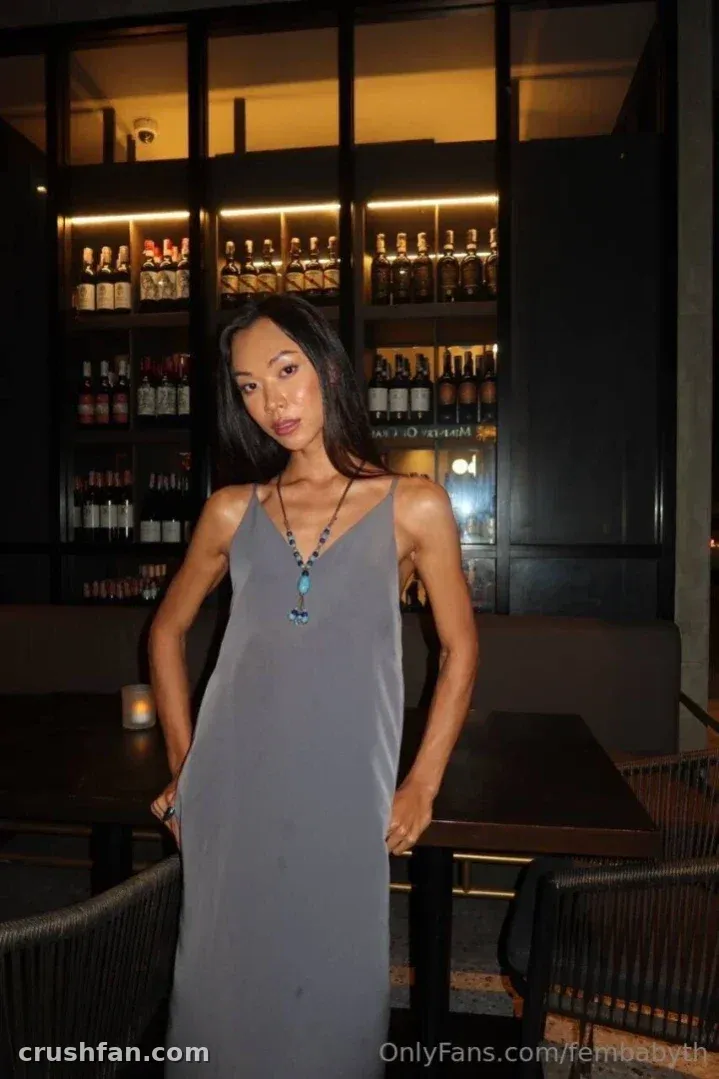 Femby in a grey slip dress standing in a dimly lit bar with bottle shelves behind her