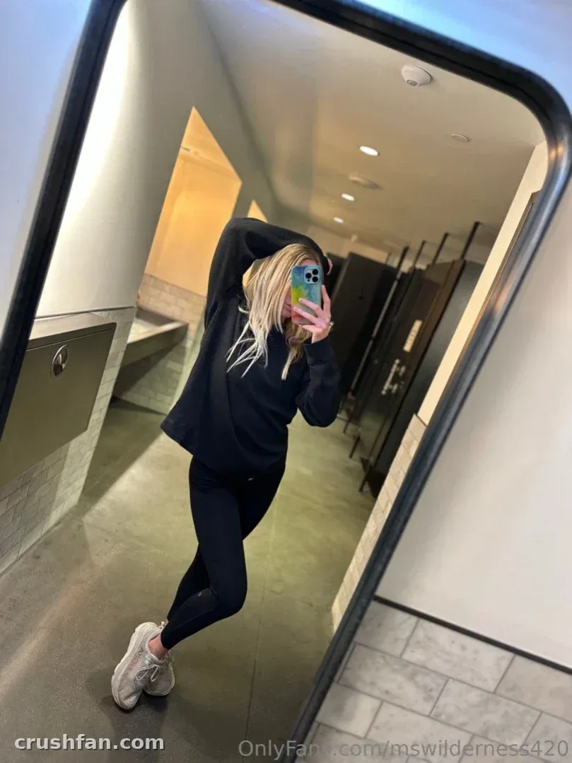 Ms Wilderness in black hoodie and leggings taking a mirror selfie in a hallway