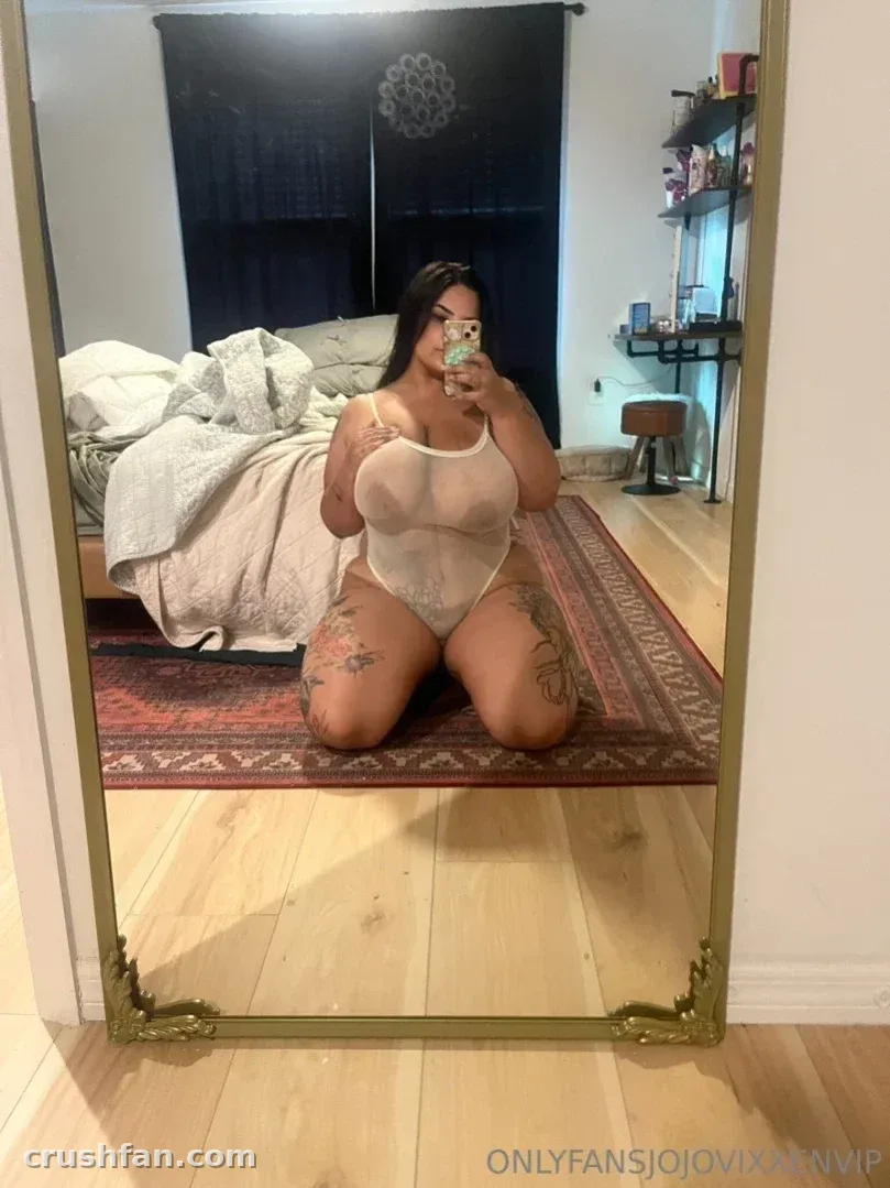 Selfie frente al espejo de una mujer de rodillas con un body claro en un dormitorio con una alfombra estampada