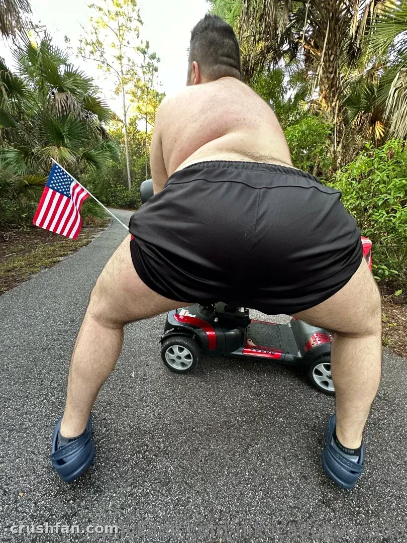 Creator from behind on a paved path with a mobility scooter, black shorts, flag and palms nearby.