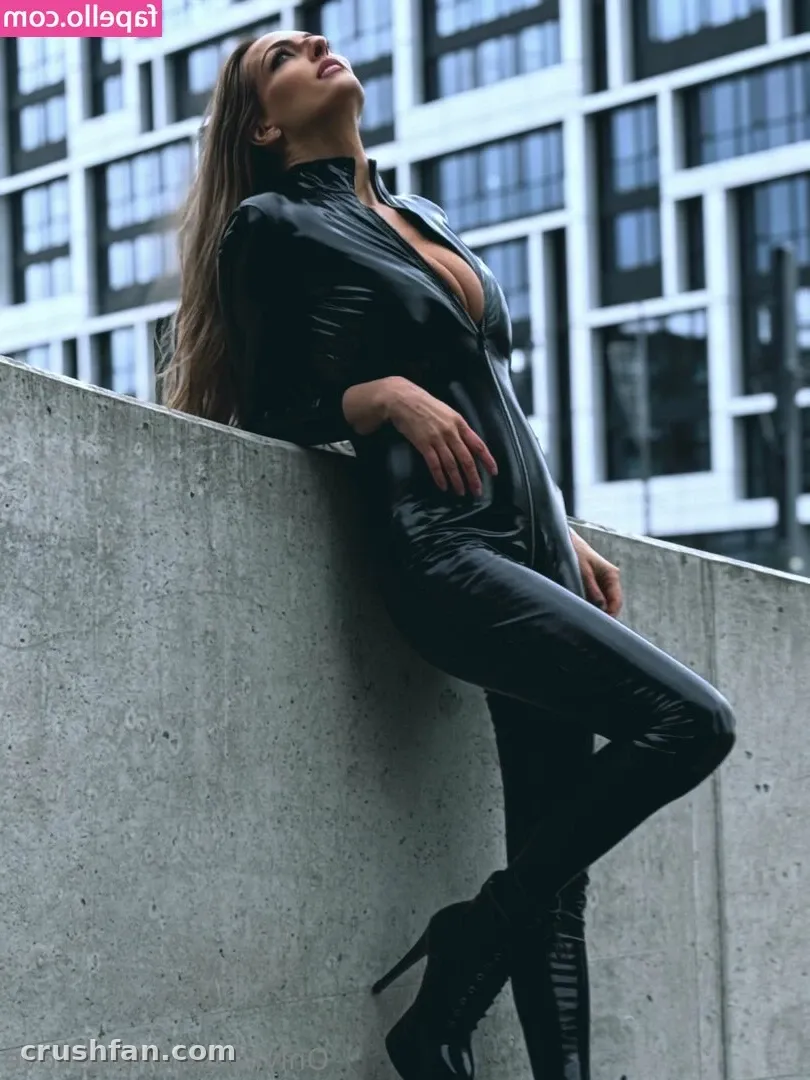 Creator in a glossy black catsuit leaning against a concrete wall with a modern building behind her