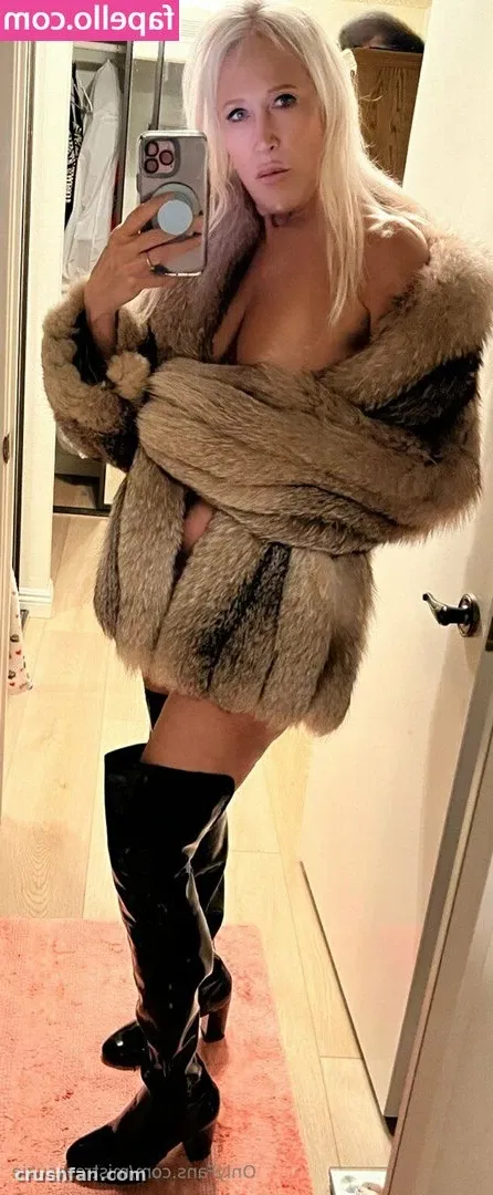 Creator in a brown fur coat and black over-the-knee boots taking a mirror selfie in a hallway