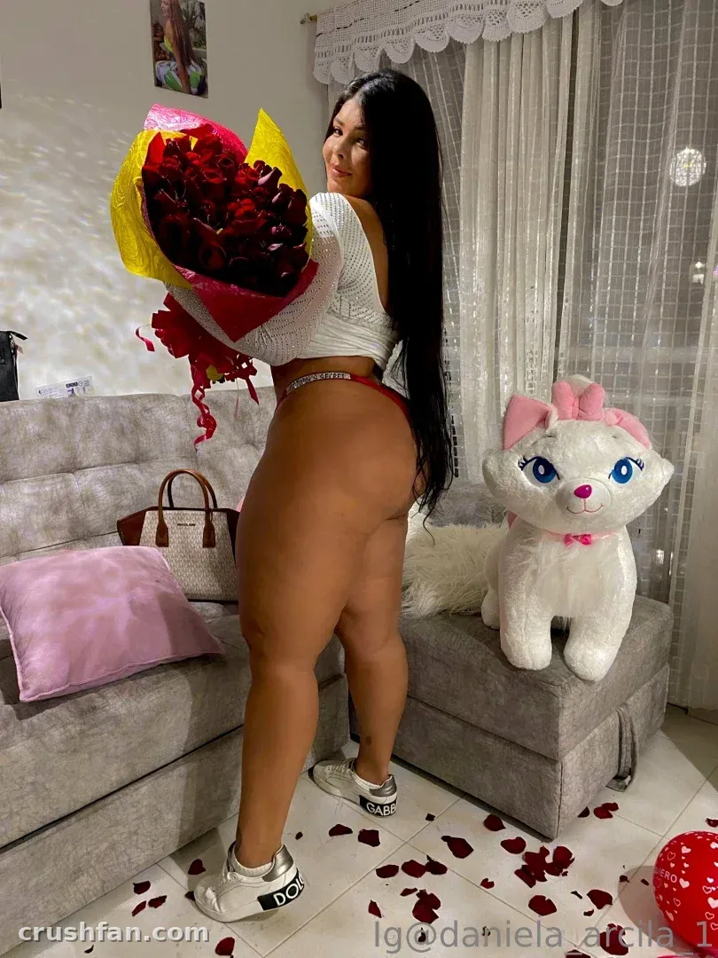 Mulher segurando um buquê de rosas vermelhas ao lado de um sofá cinza com um gato de pelúcia.