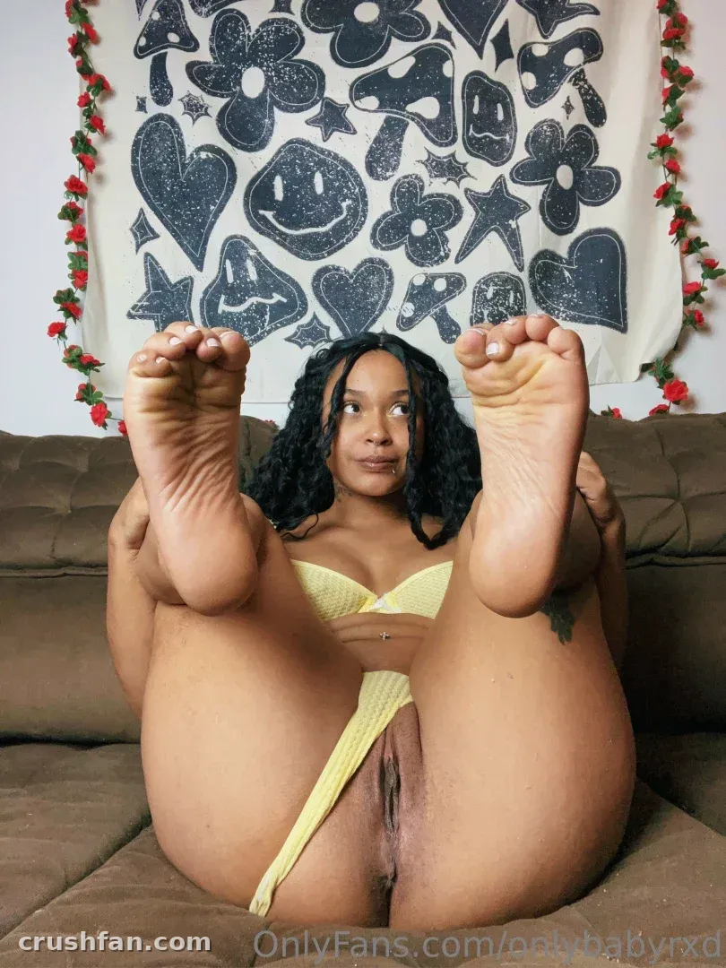 Nara on a brown sofa with bare feet raised, in front of a black and white floral tapestry