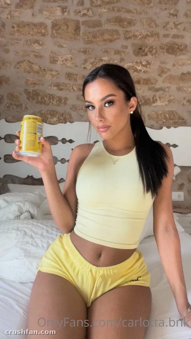 Carlotta in a yellow crop top and shorts sitting on a bed holding a yellow can.