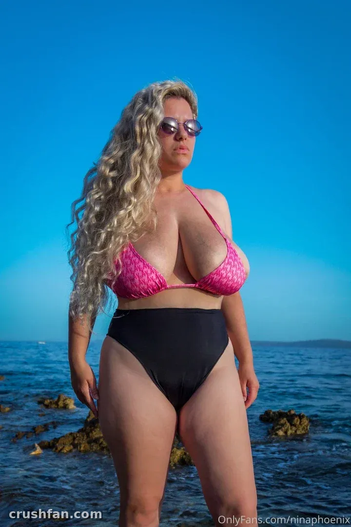 Nina Phoenix com um top de biquíni rosa e roupa de banho preta em pé à beira-mar sob um céu azul