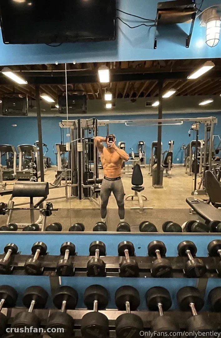 Nate tira uma selfie sem camisa no espelho em uma academia com halteres e paredes azuis.