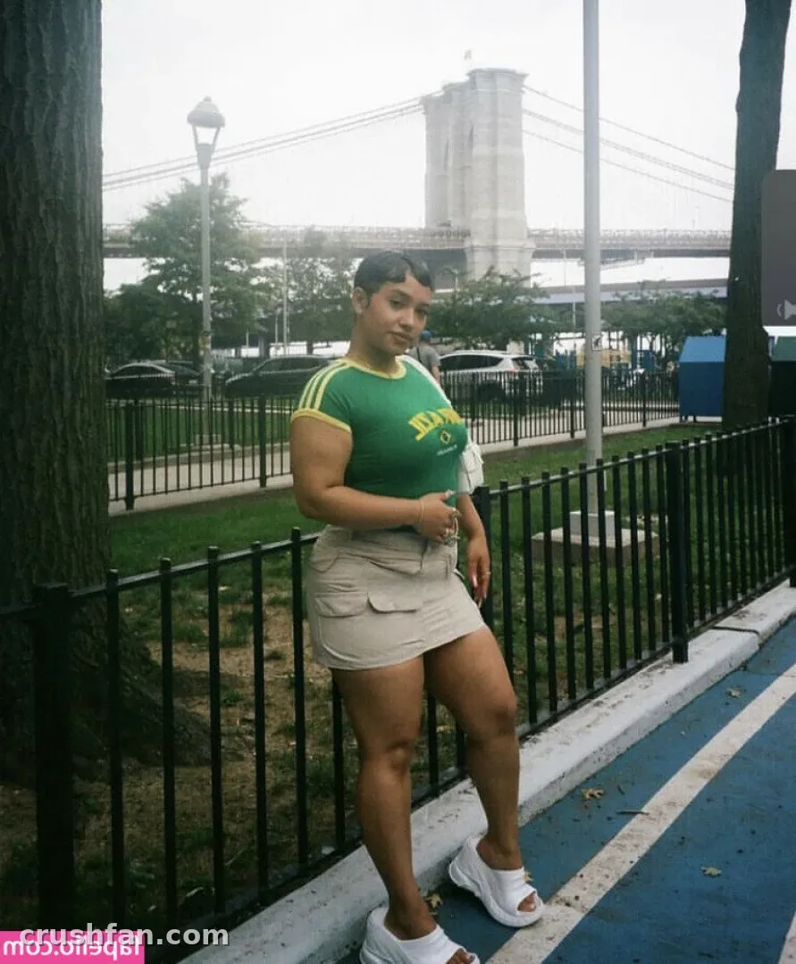 AmberThickk en haut vert et jupe beige debout près d'une clôture avec un pont derrière elle