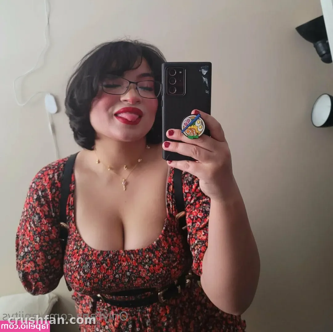 Vaniity in einem Kleid mit Blumenmuster, die ein Telefon für ein Spiegel-Selfie vor einer schlichten Wand hält