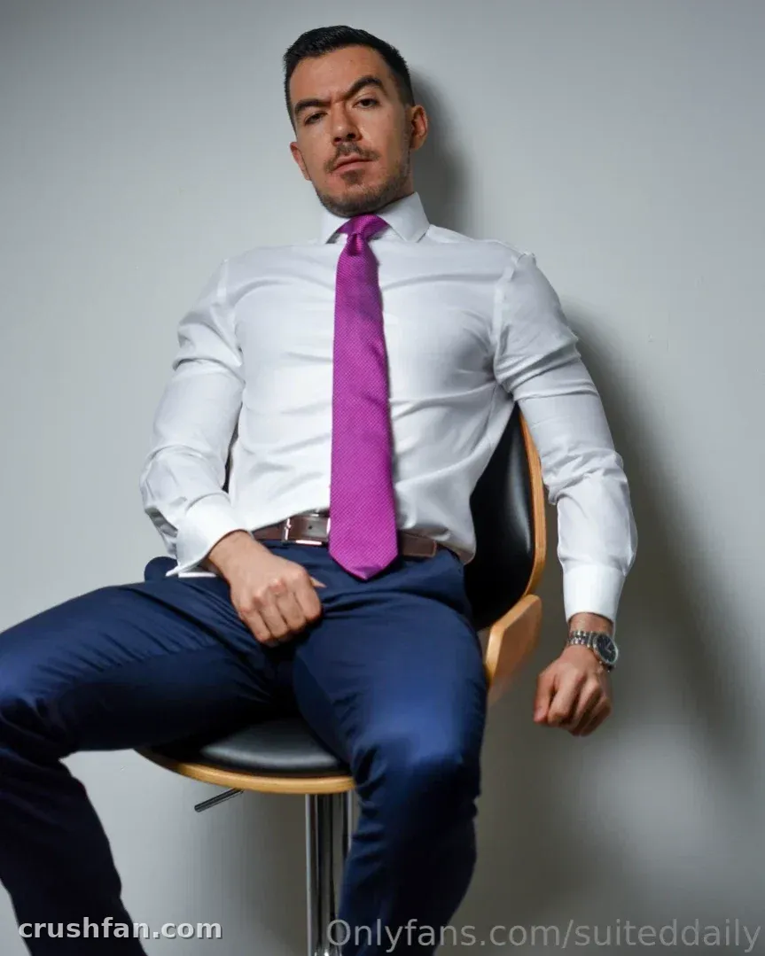 Man in a white shirt and purple tie seated on a chair against a grey wall