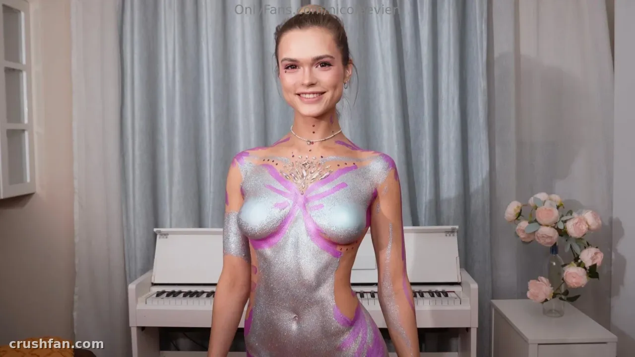 Nicole Vien with silver and pink body paint smiling in front of a white piano and grey curtains